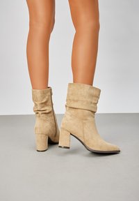 Bottes de cheville en suede beige avec un design flou, un bout pointu et un talon carré. Comprend une fermeture éclair sur le côté et une texture douce.