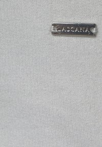 Bílý textil s jemnou strukturou, se stříbrným obdélníkovým štítkem, na kterém je napsáno "LASCANA." Celkový design je minimalistický.