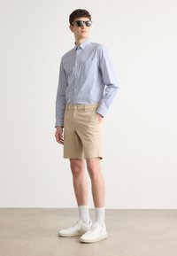 Stripet blå og hvit skjorte med knapper, beige shorts og hvite joggesko med teksturerte såler, kombinert med mørke solbriller. Enkel design.