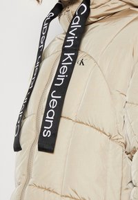 Beige pufferjakke med quiltet tekstur, der har en frontlynlås og sorte logo-remme med teksten "Calvin Klein Jeans."
