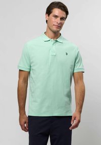 Mężczyzna w jasnoróżowej koszulce polo z krótkim rękawem i małym logo na piersi, w ciemnych spodniach, na jednolitym tle.