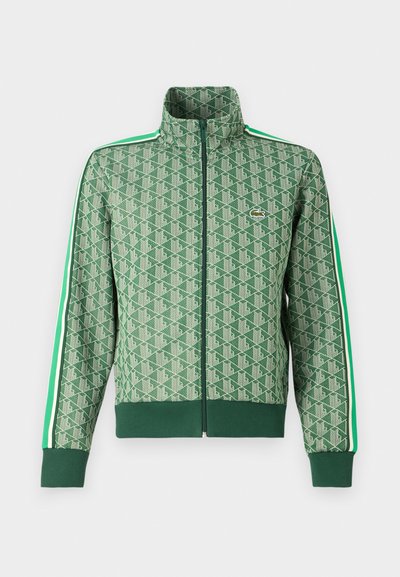 Veste de sport verte avec un motif géométrique répétitif, col montant, fermeture éclair et bandes blanches contrastantes le long des manches.
