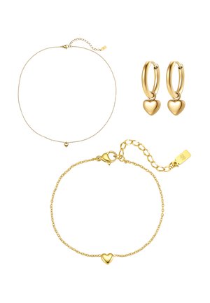 Gouden sieradenset met een delicate kettinghalsketting, armband met hartvormige hanger en oorringen met hartvormige hangers op een witte achtergrond.