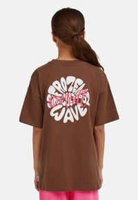 Brun oversized t-shirt med stor vit och rosa grafisk text på baksidan. Korta ärmar och en klassisk rund halsringning.