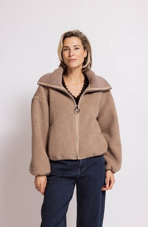 Woman with short blonde hair wearing a beige fuzzy zip-up jacket and dark blue jeans standing against a plain light background.
