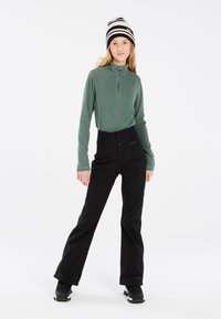 Pull en polaire vert avec col zippé, associé à un pantalon de ski noir avec une taille large et une fermeture bouton, accompagné de bottes de neige noires.