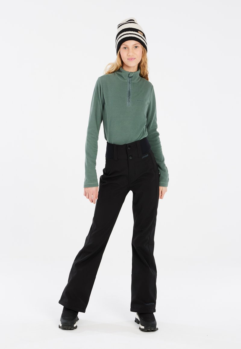 Pull en polaire vert avec col zippé, associé à un pantalon de ski noir avec une taille large et une fermeture bouton, accompagné de bottes de neige noires.