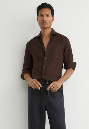 Mann trägt dunkelbraunes Hemd mit Knöpfen und hochgekrempelten Ärmeln sowie dunkle Hose, hält eine Sonnenbrille und steht vor einer weißen Wand.