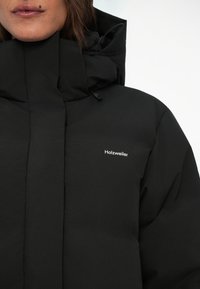 Giubbotto invernale nero con colletto alto, dotato di chiusura con zip e un piccolo logo bianco. Il materiale appare liscio e opaco.