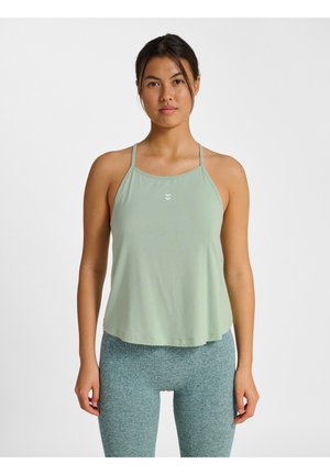 Kobieta stojąca przodem, ubrana w jasnielo zielony bezrękawnik do ćwiczeń i niebiesko-szare legginsy na jednolitym tle.