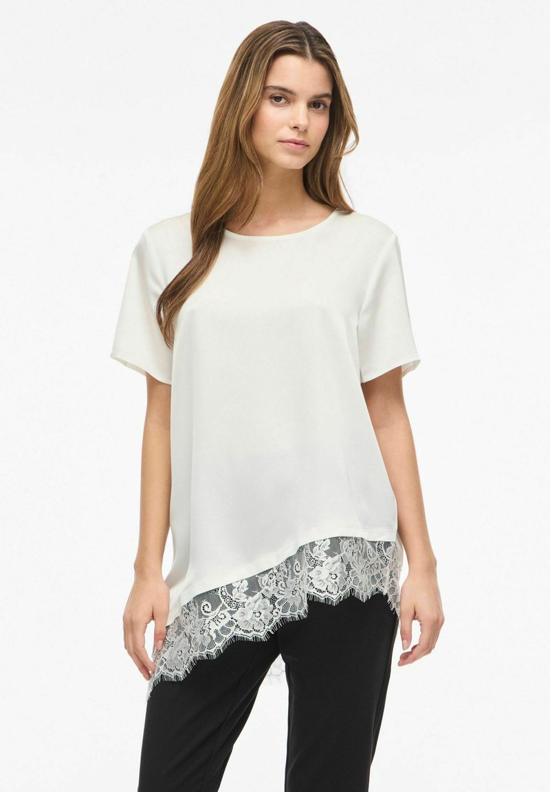Jeune femme portant un chemisier blanc ample à manches courtes avec un ourlet en dentelle asymétrique, associé à un pantalon noir, debout sur un fond uni.