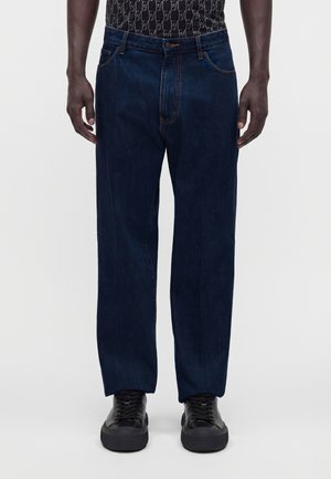Homme portant un jean droit bleu foncé, des baskets noires et une chemise noire à motifs, debout devant un fond blanc uni.