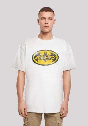 Valkoinen puuvillainen t-paita, jossa on kulunut keltainen ja musta Batman-logo, jossa on japaninkielistä tekstiä soikeassa muotoilussa. Lyhyet hihat ja pyöreä kaula-aukko.