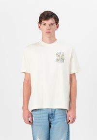 Cream cotton t-shirt with short sleeves, round neckline, and a floral graphic on the left chest area. Pairing light blue distressed jeans.