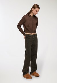 Giovane donna che indossa un hoodie corto marrone, jeans scuri larghi e scarpe slip-on beige, in piedi e guardando verso il basso su uno sfondo semplice.