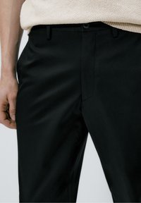 Mustat housut, joissa on sileä pinta, suora etuosa, sivutaskut ja napillinen kiinnitys. Yhdistetty vaalean sävyiseen neulepuseroon.