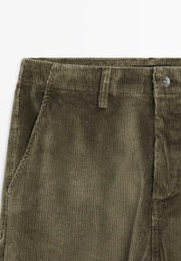 Olivenfarvede kordfløjspants med lodret ribbet tekstur, der har en frontlomme, knaplukning og bæltestropper i taljen.