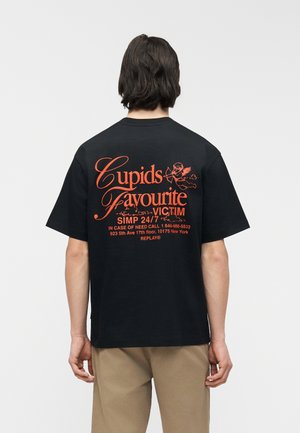 Άτομο με σκούρα μαλλιά που φοράει μαύρο T-shirt με πορτοκαλί κείμενο και γραφικό αγγέλου στο πίσω μέρος που γράφει "Cupids Favourite Victim SIMP 24/7."