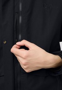 Giacca nera con chiusura a zip, texture del tessuto liscia e dettagli con bottoni. Logo visibile sul lato destro superiore. Mano che regola la zip.