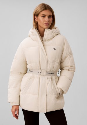 Kvinde iført en cremefarvet dunjakke med hætte, bælte i taljen med Calvin Klein-logo, hænderne i lommerne, stående mod en ensfarvet grå baggrund.