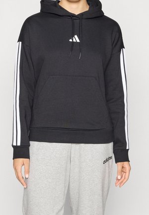 Osoba ubrana w czarną bluzę z kapturem z białymi paskami na rękawach i białym logo, w zestawie z jasnoszarymi spodniami dresowymi Adidas.