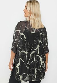 Kimono nero trasparente con motivo floreale in verde chiaro. Apertura frontale, maniche corte e spacchi laterali accentuano il design fluido.