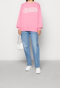 Pink sweatshirt with "Hollister California" graphic, loose fit, paired with light blue jeans and white sneakers; small white handbag as an accent.