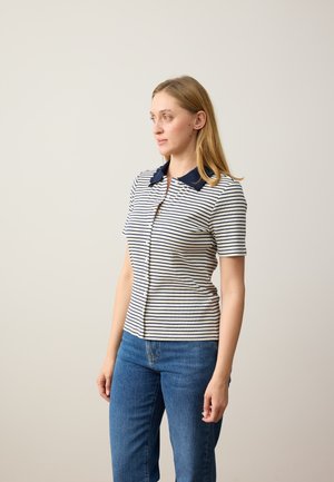 Vrouw met lichtbruin haar die een wit shirt met korte mouwen, marineblauwe strepen en kraag draagt, en een blauwe spijkerbroek, staand tegen een effen achtergrond.