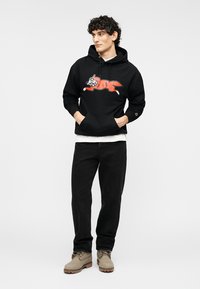 Μαύρο φούτερ με κουκούλα που διαθέτει κόκκινο γραφικό σκύλου σε στυλ καρτούν, συνδυασμένο με μαύρο παντελόνι και μπεζ μποτάκια, σε ουδέτερο φόντο.