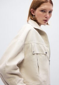 Μπεζ ζακέτα από ανθεκτικό ύφασμα, με κλείσιμο με φερμουάρ, πλαϊνές τσέπες και μινιμαλιστικό σχεδιασμό με όρθιο γιακά.