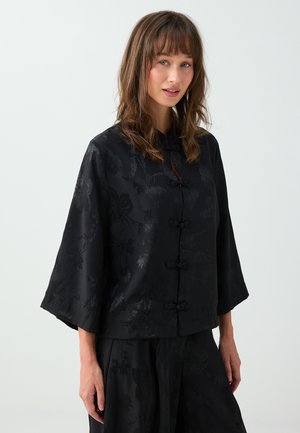 Femme aux cheveux bruns mi-longs porte un haut noir à motif floral avec trois fermetures à boutons grenouille et des manches larges.