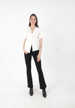 Top blanco de manga corta cruzado con un cierre floral, combinado con pantalones de campana negros y botas negras de punta en un fondo sencillo.