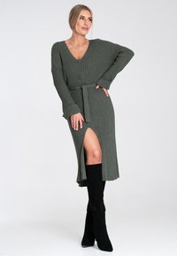 Rippiertes, olivgrünes Kleid mit V-Ausschnitt, Knopfdetails und Taillengürtel. Verfügt über einen seitlichen Schlitz und wird mit schwarzen kniehohen Stiefeln kombiniert.