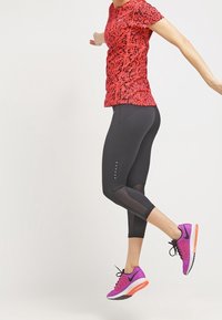 Punainen kuvioitu urheilupaita, mustat capri-legginssit verkkoyksityiskohdilla ja vaaleanpunaiset juoksukengät mustilla yksityiskohdilla ja valkoisilla pohjilla.