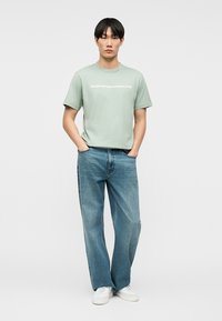 Νέος άνδρας που στέκεται με τα χέρια στις τσέπες, φορώντας ένα ανοιχτοπράσινο t-shirt Emporio Armani, άνετα μπλε τζιν και άσπρα αθλητικά παπούτσια, σε απλό φόντο.