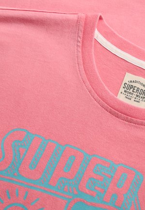 Roze T-shirt met ronde hals en turquoise "SUPERDRY" tekst, met een zichtbaar beige merkembleem aan de binnenzijde van de kraag.