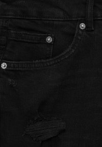 Gros plan d'une poche de jean en denim noir avec des rivets métalliques et une petite déchirure usée sous la poche.