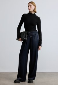 Melnas, cieši piegulošas, garām piedurknēm krekls ar augstu apkakli, kombinēts ar tumši ziliem platajiem svārkiem ar elastīgu jostu. Modeles rokās melna rokassoma.