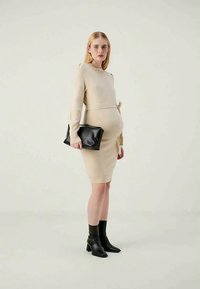 Adīta bežīga kleita ar garām piedurknēm un sasaistes detaļu, apvienota ar melnu somu un potītes zābakiem, izceļas ar pieguļošu siluetu.