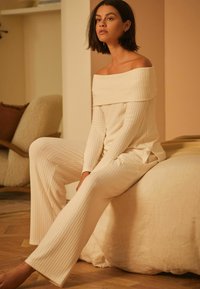 Off-shoulder ribbel-cremekleurige top gecombineerd met bijpassende ribbelbroek. De stof lijkt zacht met een textuurafwerking. Eenvoudig, elegant ontwerp.