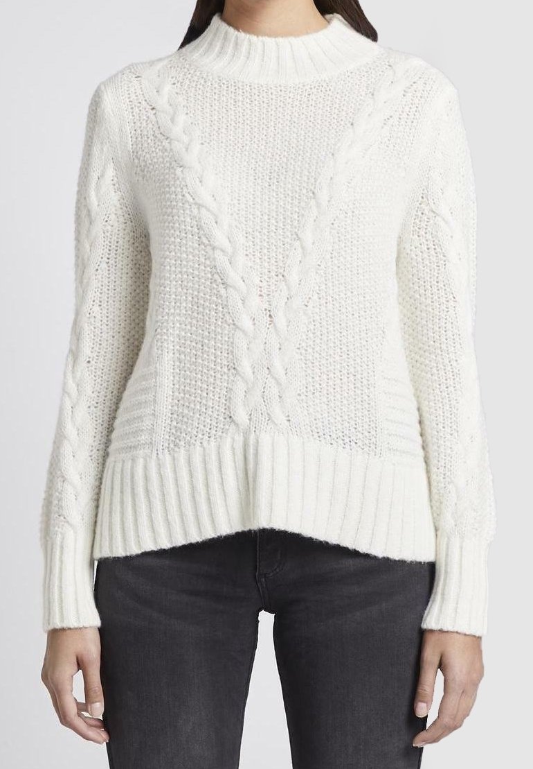 Femme portant un pull blanc en maille torsadée avec col, poignets et ourlet côtelés, associé à un jean noir, debout devant un fond uni.