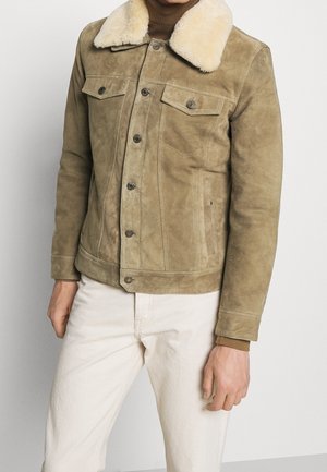 Homme portant une veste en daim beige avec un col en shearling blanc, un col roulé marron et un pantalon écru sur un fond uni.