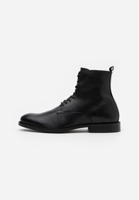 Botas de tobillo de cuero negro con un acabado suave, punta redonda, cordones en la parte frontal y una pequeña asa en la parte posterior. Suela de goma negra sólida.