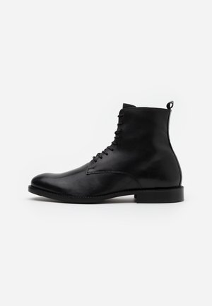 Bottines en cuir noir avec une finition lisse, bout arrondi, fermeture à lacets à l'avant et une petite tirette à l'arrière. Semelle en caoutchouc noir uniforme.