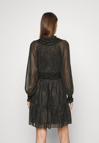 Femme aux longs cheveux bruns portant une robe noire transparente à pois blancs, manches longues, col haut et ourlet à volants, debout de dos.