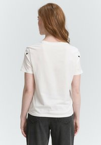 Biały bawełnowy t-shirt z krótkim rękawem, z małym czarnym haftem w kształcie serca na ramionach. Prosty, okrągły dekolt i luźny krój.