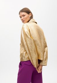 Metalni zlatni sako s teksturiranim finišom, s ovratnikom i dva prednja džepa, nosi se preko ljubičastih hlača.