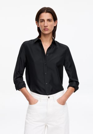 Mujer con cabello castaño oscuro que lleva una camisa negra abotonada con las mangas remangadas y pantalones blancos, manos en los bolsillos, fondo neutro.