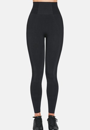 Kobieta ubrana w czarne, bezszwowe legginsy z wysokim stanem i czarne buty sportowe, stojąca na tle jednolitego jasnego tła.