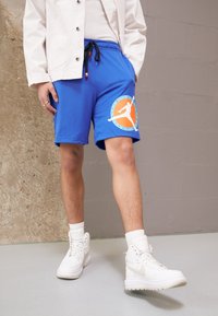 Blue athletic shorts with black drawstring, mesh texture, featuring a large orange and white logo. Paired with white sneakers and socks.